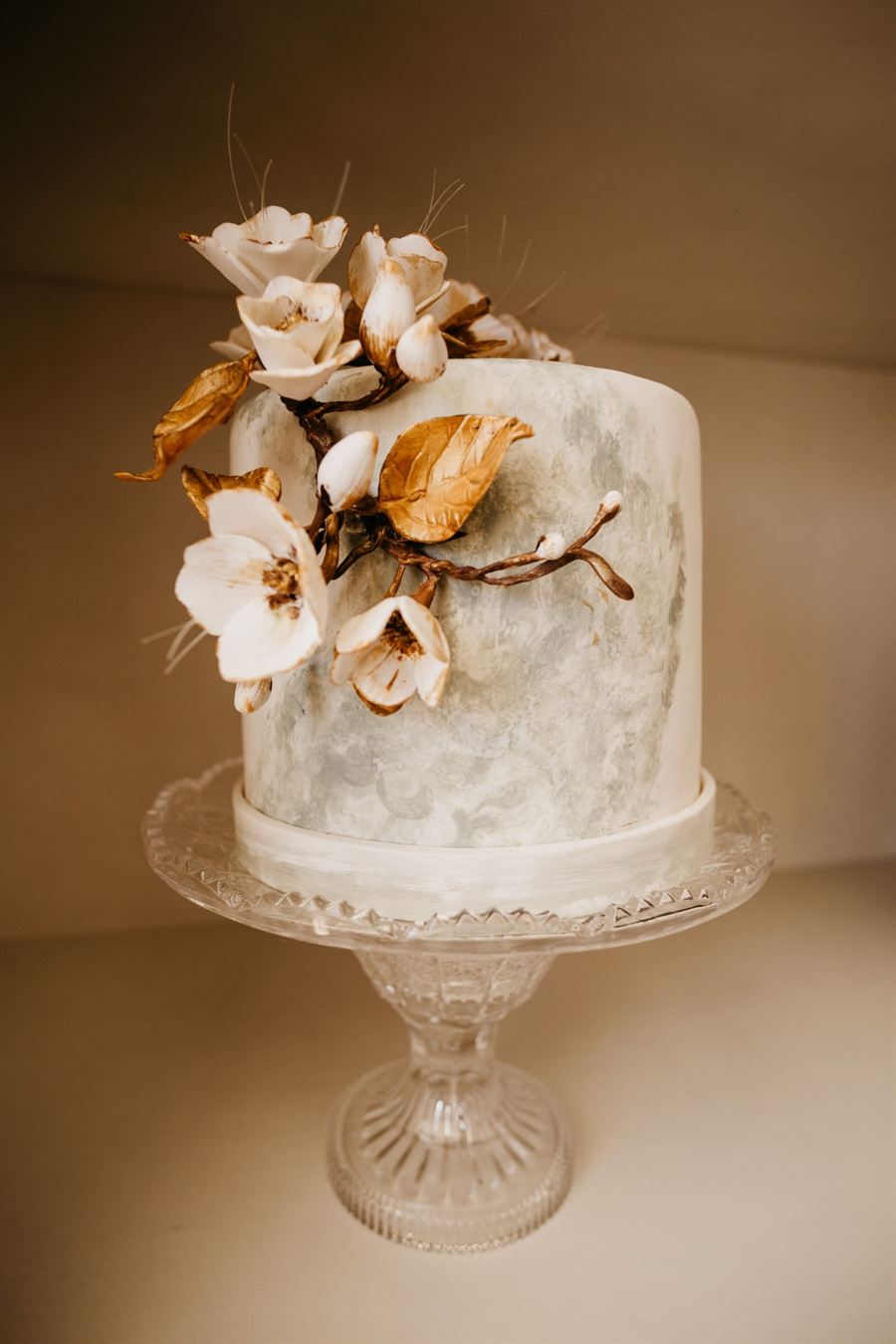Bolo de casamento branco com flores de açúcar e folhas douradas num prato de cristal