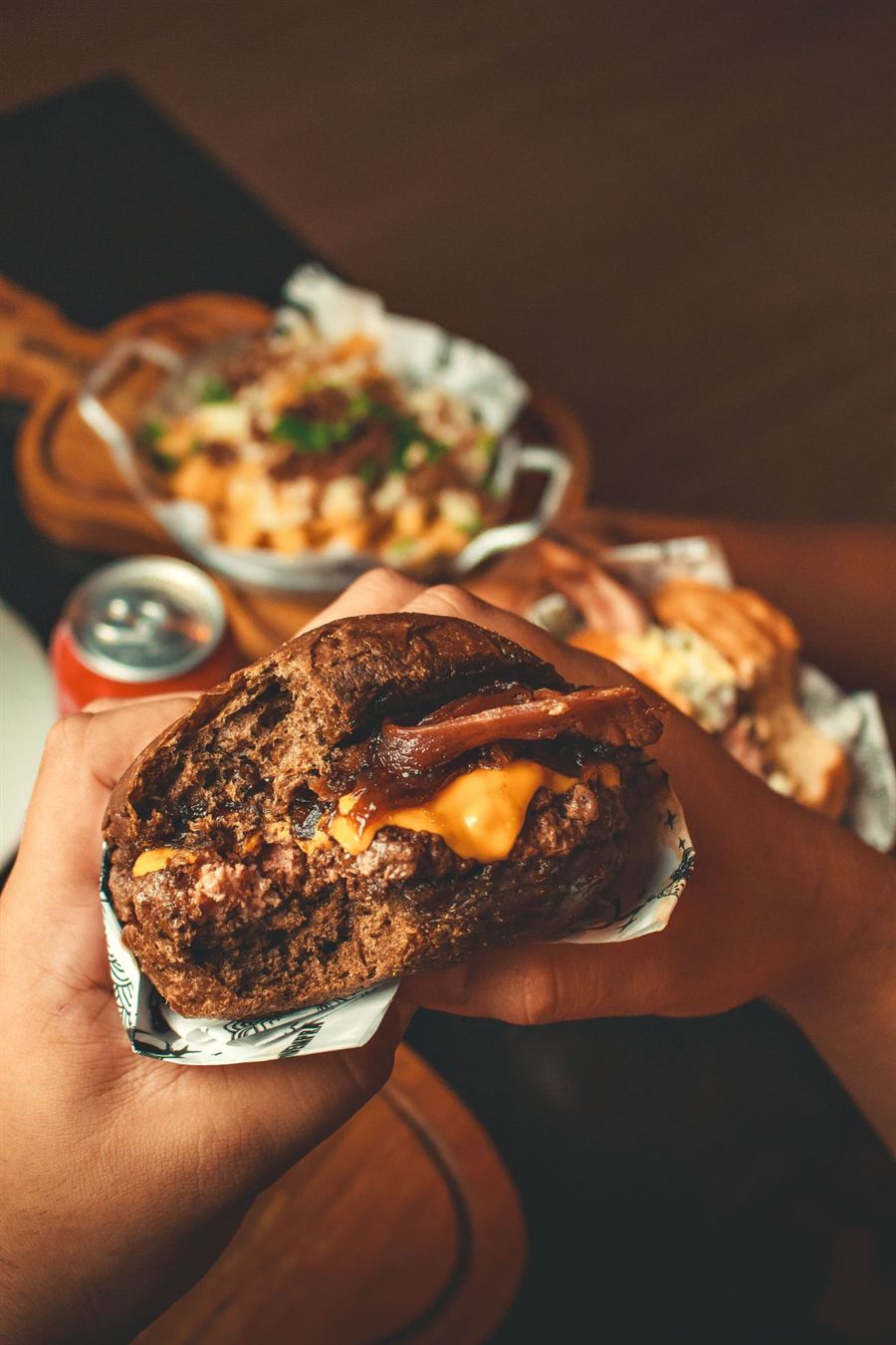 Mão segurando um burger de pão escuro com cheddar, bacon e cebola caramelizada