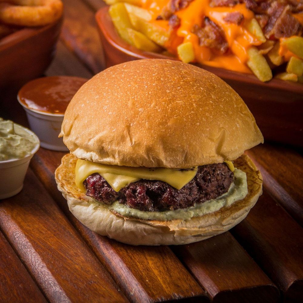 Hambúrguer com queijo sobre tábua de madeira, com batata com cheddar e bacon e molhos ao fundo
